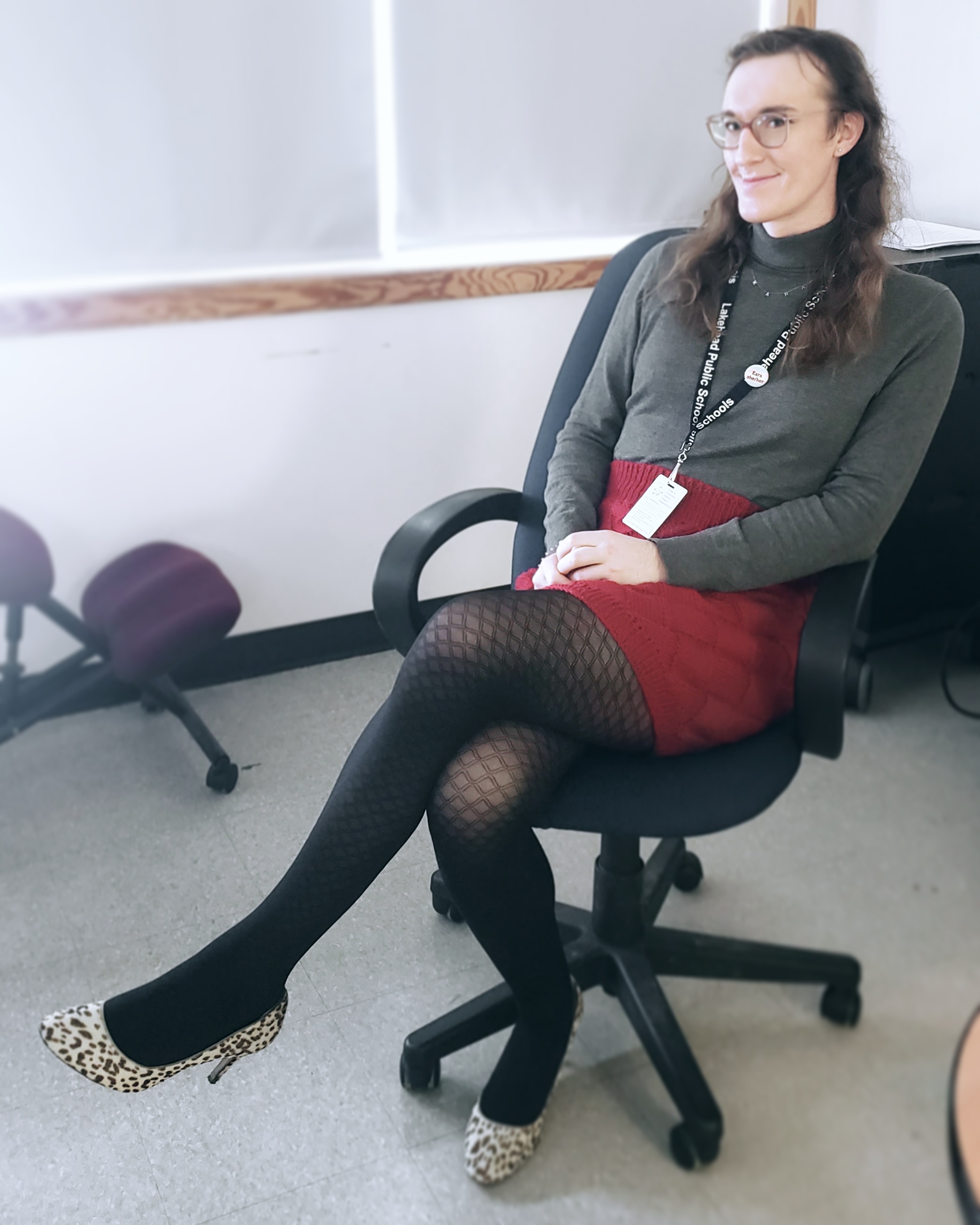 I'm sitting with my legs crossed in an office chair. I'm wearing a knitted red skirt and grey turtleneck with diamond-pattern tights and leopard ankle booties. My hair is down, and I am smiling.