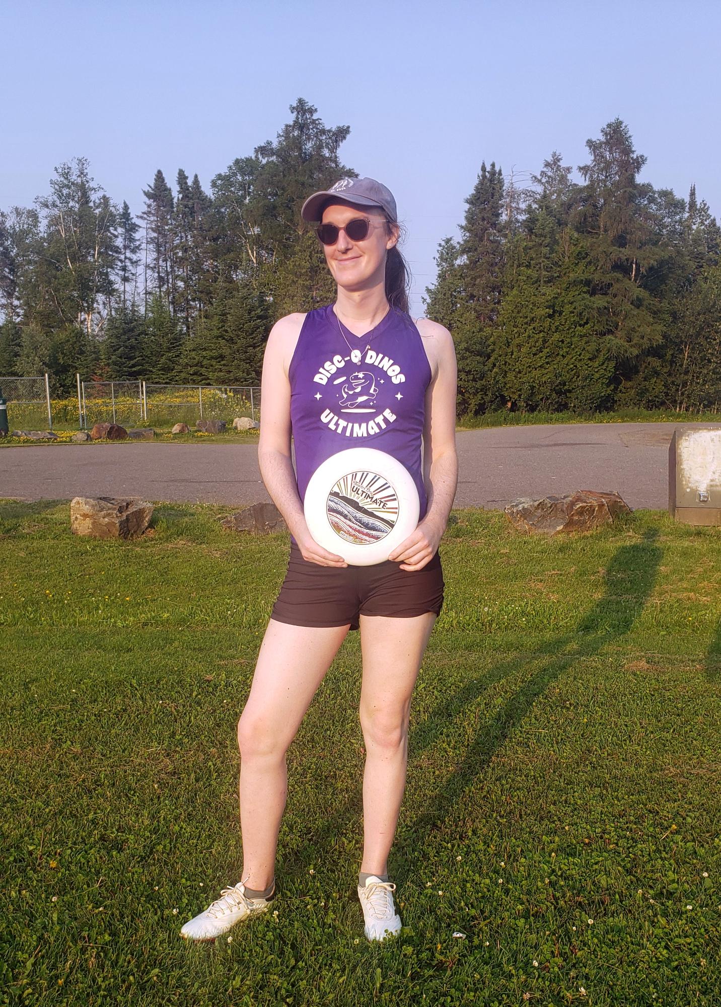 Photo of me standing outdoors. I'm wearing black bicycle shorts and a purple tank top with a white logo that says 'Disc-o-Dinos' on it with a picture of a dinosaur. My hair is in a ponytail under a baseball cap, and I have sunglasses on. I'm smiling. I'm holding a white frisbee that says 'Thunder Bay Ultimate.'